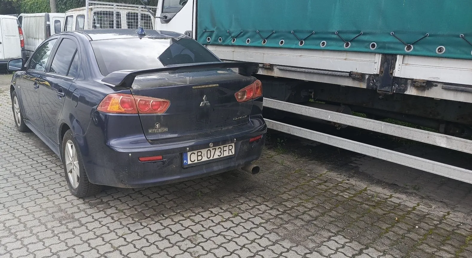 Granatowy samochód osobowy marki Mitsubishi Lancer zaparkowany tyłem na kostce brukowej obok białej ciężarówki z zieloną plandeką. Samochód posiada spojler na klapie bagażnika oraz polską tablicę rejestracyjną o numerze CB 073FR.