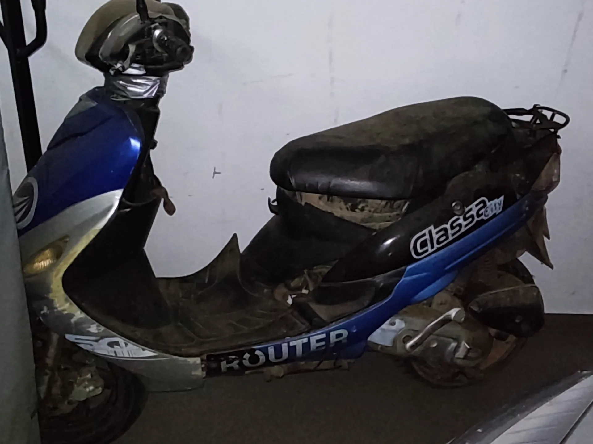 Niebiesko-srebrny skuter w stanie zużycia z czarnym, zużytym siedzeniem. Na boku widoczne napisy 'Classa City' oraz 'ROUTER'. Na kierownicy umieszczony kask. Tło stanowi jasna ściana i podłoga.