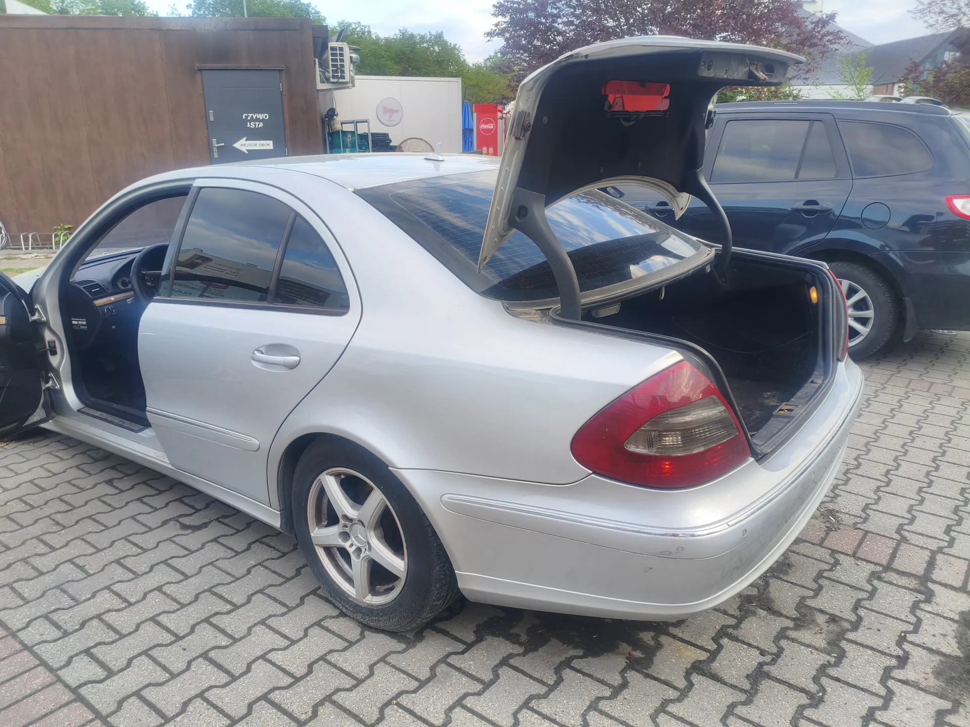 Srebrny samochód osobowy typu sedan z otwartym bagażnikiem i drzwiami kierowcy, zaparkowany na brukowanym placu. W tle ciemnoszary SUV, drewniana budowla z drzwiami z napisem "CZYWO JSTA" oraz strzałką w lewo i napisem "WJŚCIE OBOK", czerwony automat z logo Coca-Cola oraz drzewa. Widoczne są również fragmenty innych budynków i niebo.
