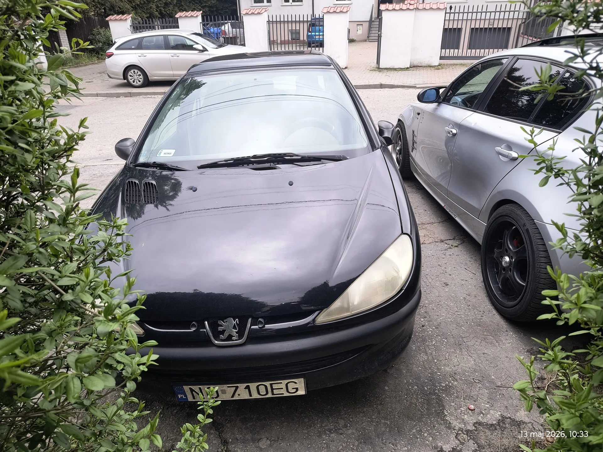 Przód czarnego samochodu marki Peugeot z tablicą rejestracyjną ND 710EG (PL) na pierwszym planie. Po prawej stronie srebrny samochód. W tle srebrny samochód zaparkowany na ulicy, biały budynek z czarnym ogrodzeniem i zielonymi krzewami po obu stronach.
