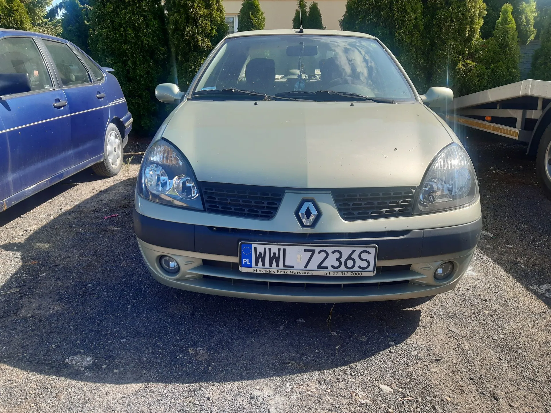 Zdjęcie przodu jasnego, złoto-beżowego samochodu marki Renault zaparkowanego na żwirowym podłożu. Samochód ma polską tablicę rejestracyjną o numerze WWL 7236S. Po lewej stronie widoczny jest fragment niebieskiego samochodu, a w tle zielone krzewy i fragment białej przyczepy po prawej stronie.