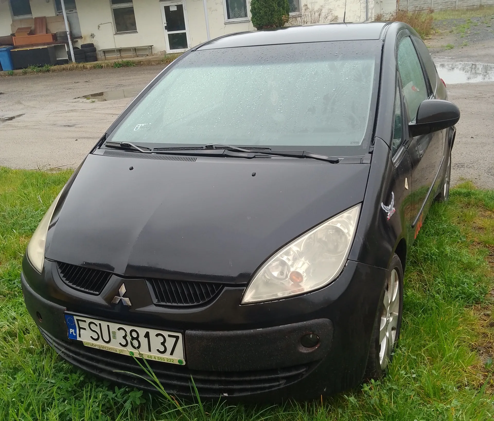 Czarny samochód Mitsubishi z tablicą rejestracyjną FSU 38137, zaparkowany na trawie. Widoczne są przednie światła, logo marki na masce, boczne lustro i naklejka z orłem na drzwiach. W tle budynek z oknami, stos opon i pojemniki. Na tablicy rejestracyjnej widoczny napis PL oraz zielony napis z numerem telefonu 68 4 555 222. Trawa i kałuże wskazują na niedawny deszcz.