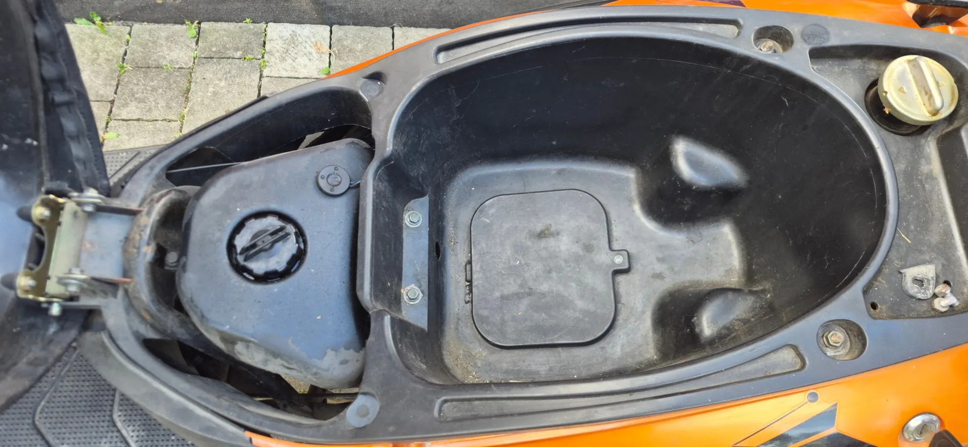 Widok z góry na otwarty schowek pod siedzeniem pomarańczowego skutera. Wnętrze schowka jest wykonane z czarnego plastiku i zawiera wgłębienia oraz prostokątną klapkę na dnie. Po lewej stronie widoczny jest czarny zbiornik z zakrętką, a po prawej żółta zakrętka wlewu paliwa.