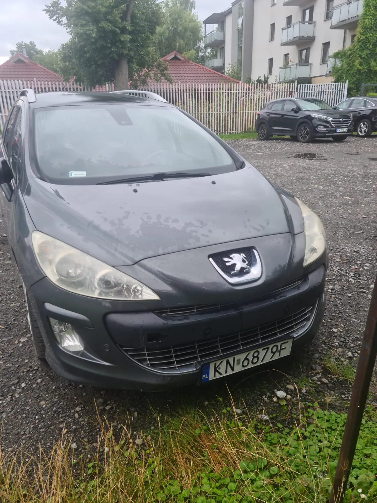 Ciemnoszary samochód marki Peugeot z tablicą rejestracyjną KN 6879F (PL) zaparkowany na żwirowym placu. W tle widoczne są czarne samochody, biały płot, budynki z czerwonymi dachami, drzewa oraz pochmurne niebo. Na przedniej szybie samochodu znajduje się naklejka.