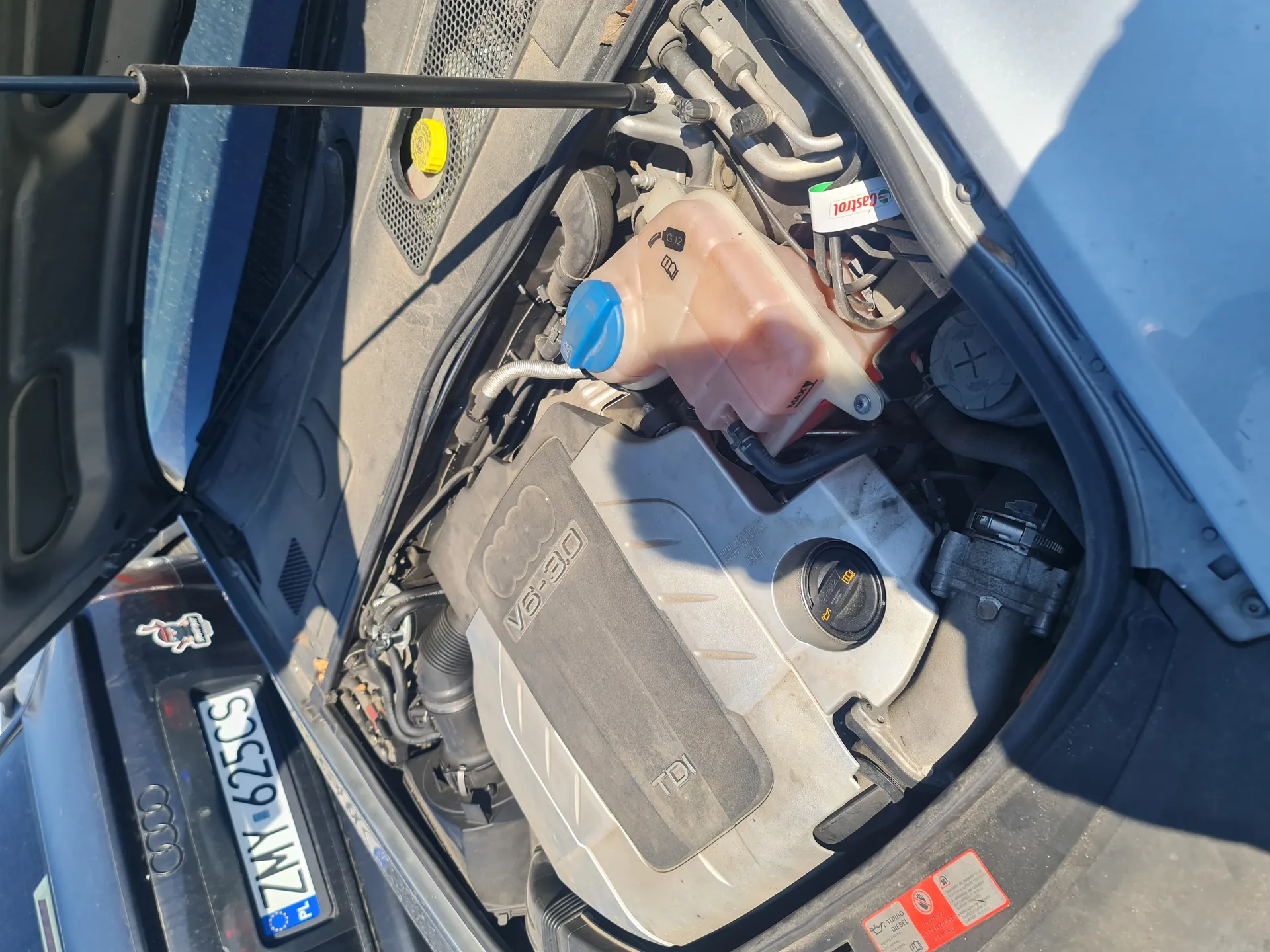 Widok otwartej tylnej części silnika samochodu marki Audi. W centrum znajduje się pokrywa silnika z napisem V6-3.0 TDI. Po lewej stronie widoczna tablica rejestracyjna ZMY 625CS z niebieskim pasem PL oraz logo Audi. W silniku widoczne są elementy takie jak zbiornik chłodniczy z niebieską pokrywką, żółta pokrywka oleju, przewody i etykieta Castrol. Na dole po prawej stronie znajdują się czerwone naklejki ostrzegawcze. Otwarta klapa silnika i fragment przedniej szyby są widoczne po lewej stronie obrazu.