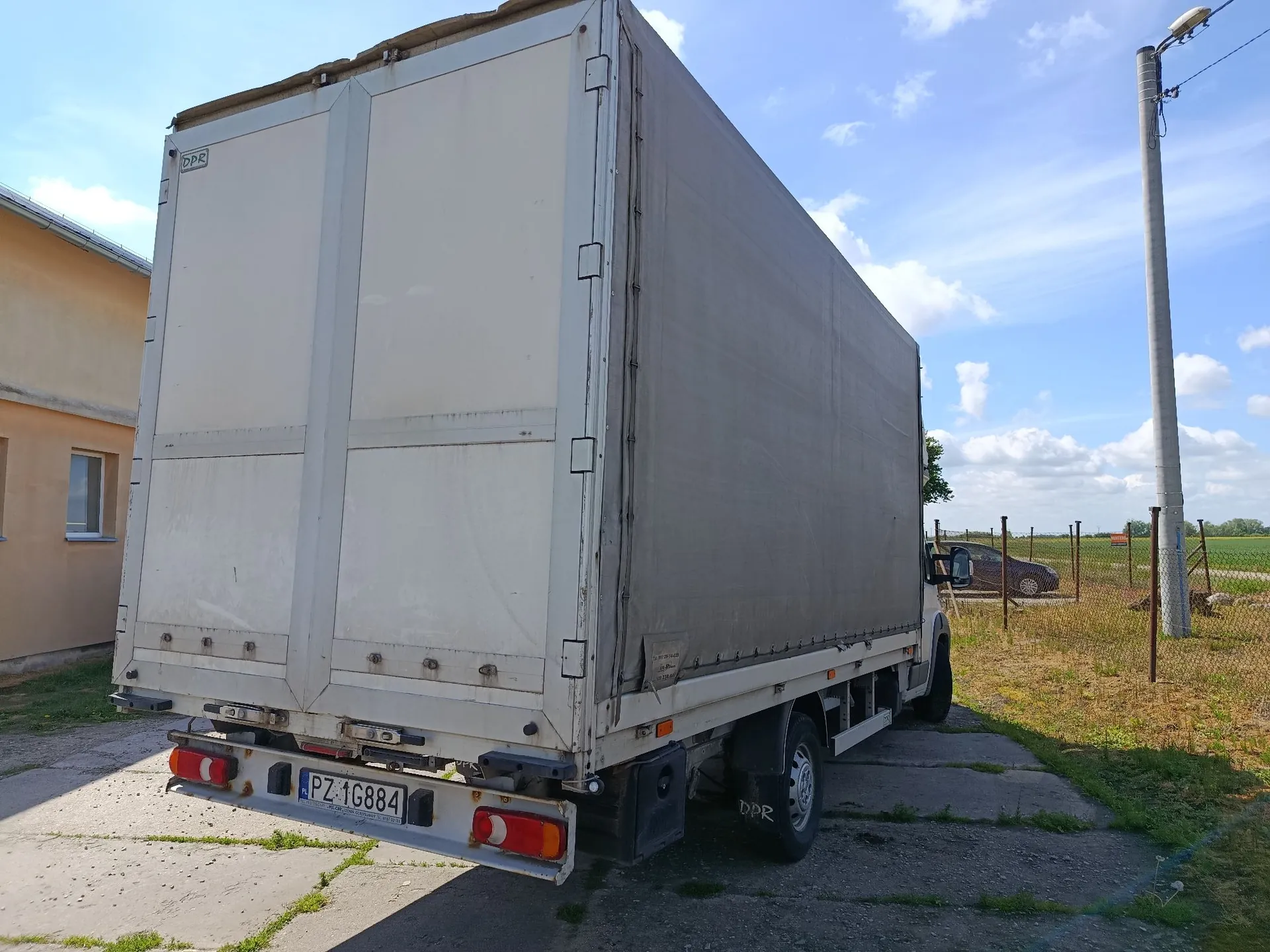 Biała ciężarówka z zabudową furgonową zaparkowana na betonowym podjeździe. Widoczny jest tył pojazdu z tablicą rejestracyjną PZ 16884 oraz tylne światła. Po prawej stronie znajduje się betonowy słup oświetleniowy, a w tle widać zielone pole i brązowy samochód osobowy pod jasnym, lekko zachmurzonym niebem.