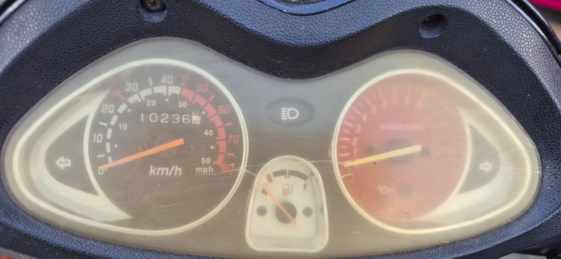 Zbliżenie na panel wskaźników skutera. Po lewej stronie znajduje się prędkościomierz z dwiema skalami: w km/h (od 0 do 80) oraz w mph (od 0 do 50). Wskazówka prędkościomierza wskazuje 0 km/h, a licznik przebiegu pokazuje wartość 10236. Nad prędkościomierzem znajduje się wskaźnik poziomu paliwa. Po prawej stronie znajduje się obrotomierz z oznaczeniem x1000 r/min. Pomiędzy wskaźnikami widoczna jest kontrolka świateł. Całość jest zamknięta w plastikowej, lekko zmatowiałej obudowie.