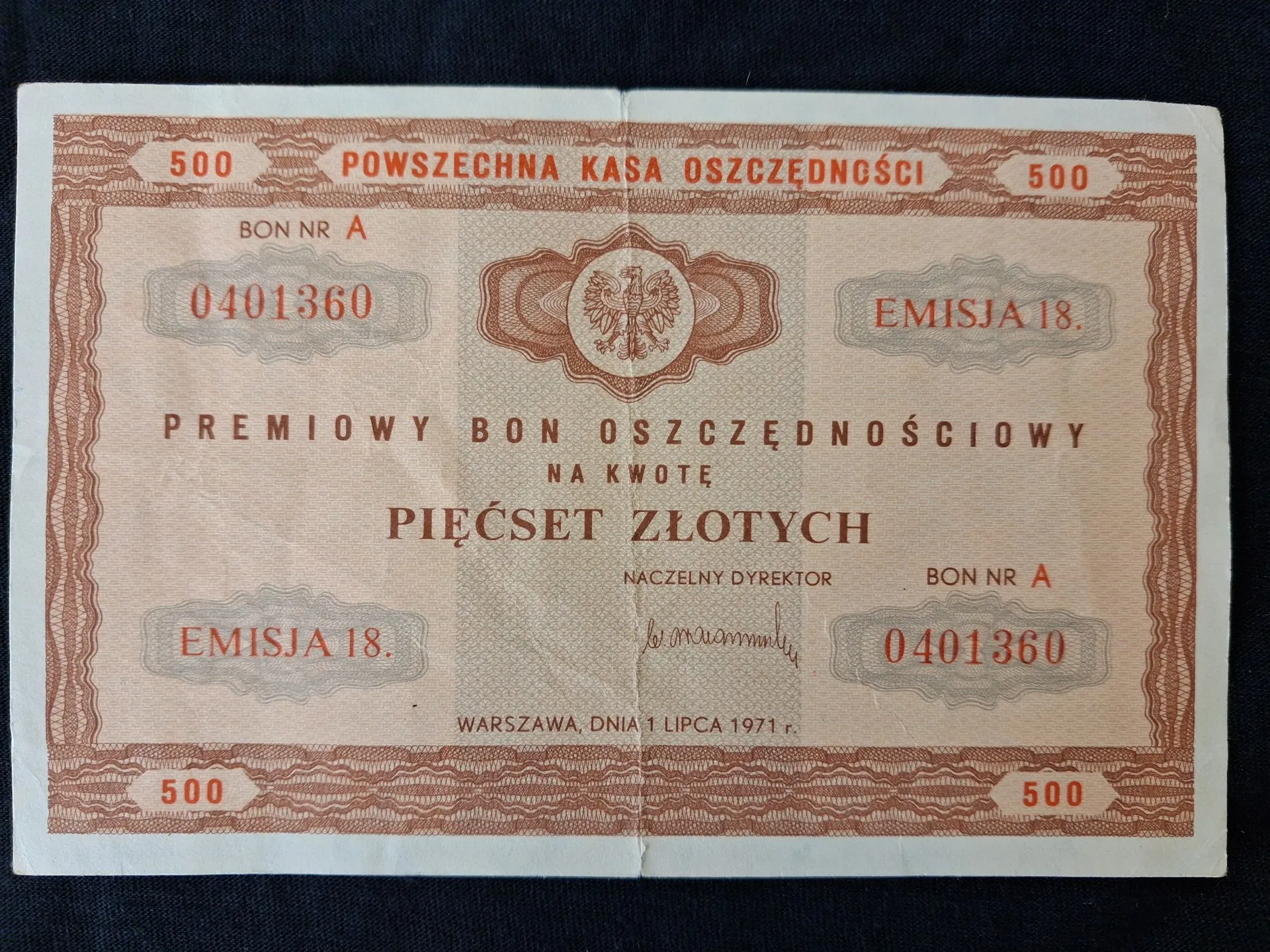 Zdjęcie starego, pomarańczowo-brązowego dokumentu finansowego. Na górze widnieje napis 'POWSZECHNA KASA OSZCZĘDNOŚCI'. W centrum znajduje się napis 'PREMIOWY BON OSZCZĘDNOŚCIOWY NA KWOTĘ PIĘĆSET ZŁOTYCH'. Dokument zawiera numer 'BON NR A 0401360' powtórzony w dwóch miejscach oraz informację 'EMISJA 18.'. W środku górnej części znajduje się godło Polski. Na dole widnieje podpis Naczelnego Dyrektora oraz data 'WARSZAWA, DNIA 1 LIPCA 1971 r.'. W czterech rogach widnieje liczba '500'.