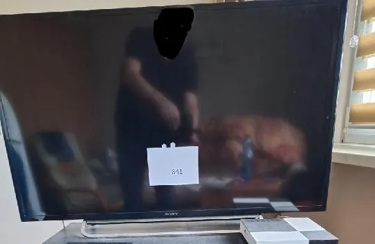 Czarny ekran wyłączonego telewizora marki Sony. Na ekranie widoczne jest odbicie osoby stojącej przed urządzeniem oraz fragmenty pomieszczenia. Na środku ekranu przyklejona jest mała, biała kartka z napisem '311'. Telewizor stoi na srebrnej podstawce, a obok niego znajduje się białe urządzenie elektroniczne.