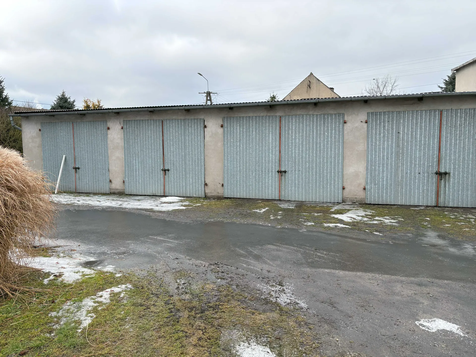 Rząd czterech garaży z metalowymi, szarymi, pionowo prążkowanymi drzwiami z czerwonymi uchwytami. Budynek o betonowej elewacji z płaskim dachem. Przed garażami nawierzchnia asfaltowa z resztkami śniegu i plamami trawy. Po lewej stronie duży stóg słomy. W tle drzewa, latarnia uliczna, słup energetyczny oraz dom z trójkątnym dachem. Niebo zachmurzone.
