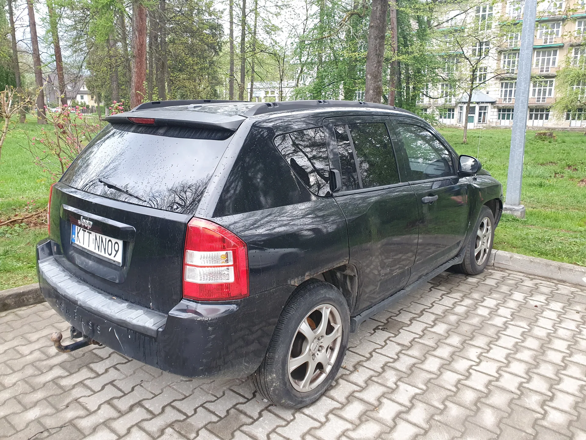 Czarny samochód typu SUV marki Jeep zaparkowany na brukowanej powierzchni. Widok z tyłu i boku. Na tablicy rejestracyjnej widoczny numer KTT NN09. Samochód ma wilgotną karoserię, co sugeruje niedawny deszcz. W tle zielona trawa, drzewa oraz budynek z wieloma oknami. Na tylnym zderzaku widoczny holownik.