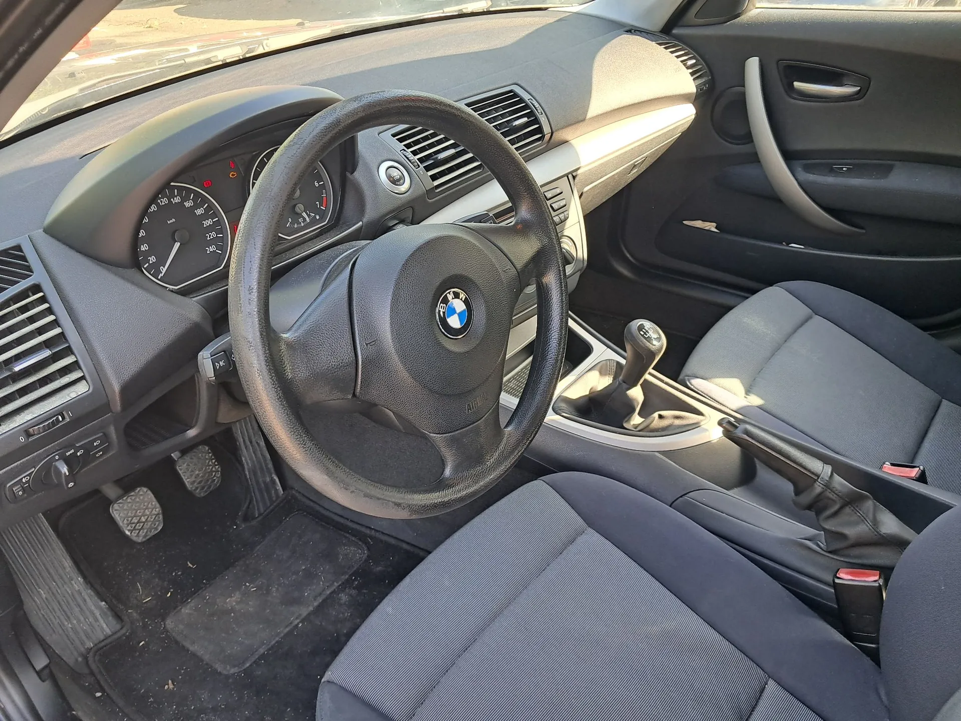 Wnętrze samochodu marki BMW z perspektywy kierowcy. Widoczna jest czarna kierownica z logo BMW w centrum, deska rozdzielcza z analogowymi wskaźnikami prędkości i obrotomierzem, lewy fotel oraz konsola środkowa z drążkiem zmiany biegów i hamulcem ręcznym. Pod nogami znajdują się pedały i czarna mata.