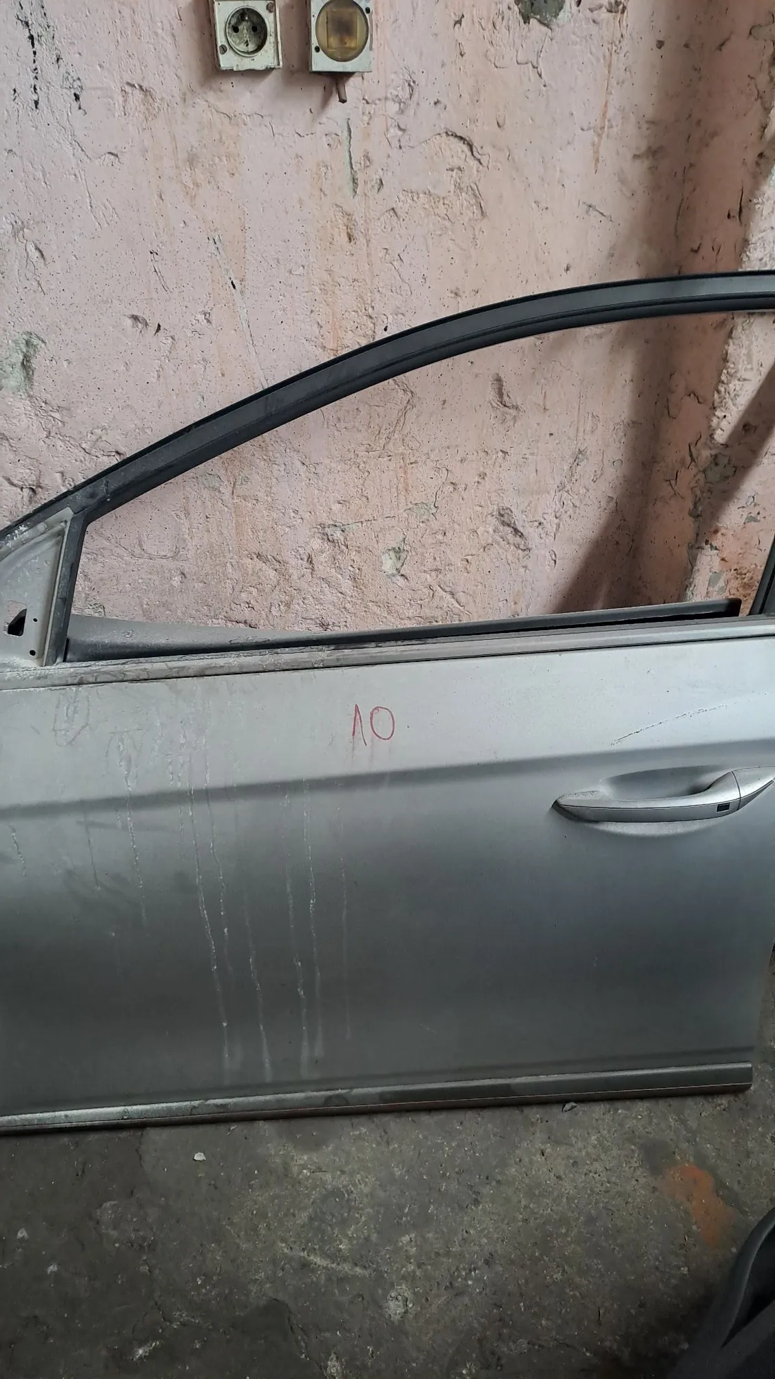 Srebrne drzwi samochodowe leżące na betonowej podłodze obok różowej, tynkowanej ściany. Na drzwiach widoczna jest klamka oraz czerwony napis 'OV'.
