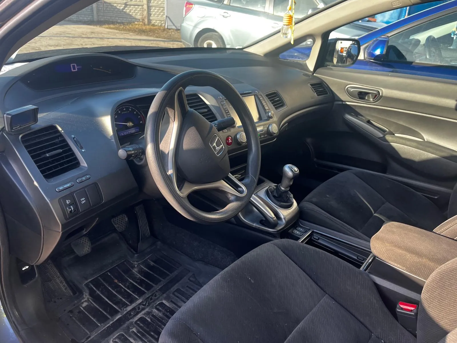 Wnętrze samochodu marki Honda z widocznym kierownicą z logo Hondy, deska rozdzielcza z licznikiem prędkości pokazującym 0 km/h, manualną skrzynią biegów, czarnymi siedzeniami tapicerowanymi tkaniną oraz dywanikami podłogowymi z napisem "Civic". W tle przez przednią szybę widoczne są zaparkowane samochody i budynek. Na lusterku wstecznym wiszący przedmiot.