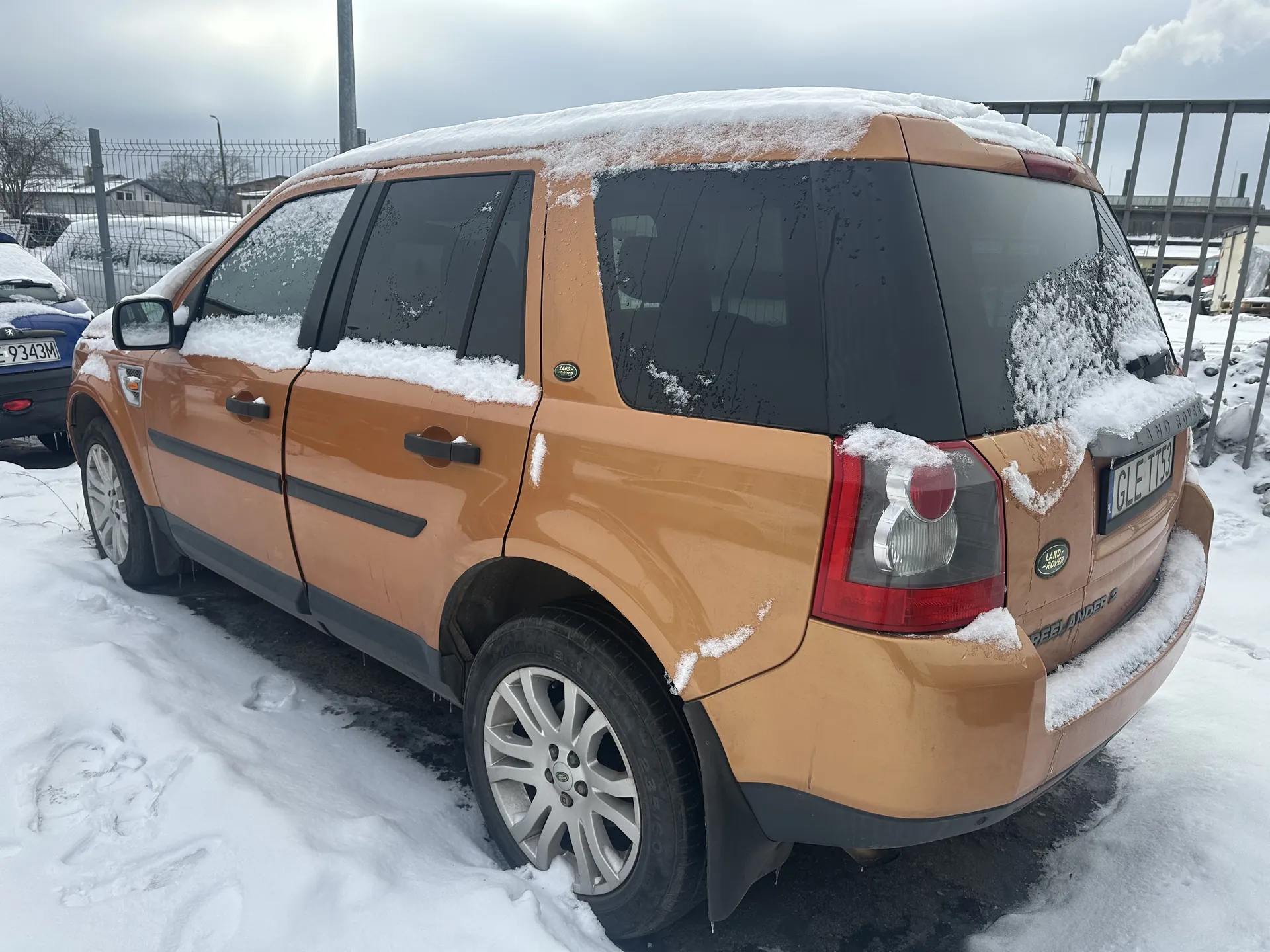 Brązowy SUV marki Land Rover Freelander 2 pokryty śniegiem, widok z tyłu i boku. Na dachu, szybach i nadwoziu widoczne śnieg. Tablica rejestracyjna GLE TT53. W tle niebieski samochód z tablicą 9343M, ogrodzenie metalowe, budynki i zasneżony teren. Niebo zachmurzone, na horyzoncie komin z dymem.