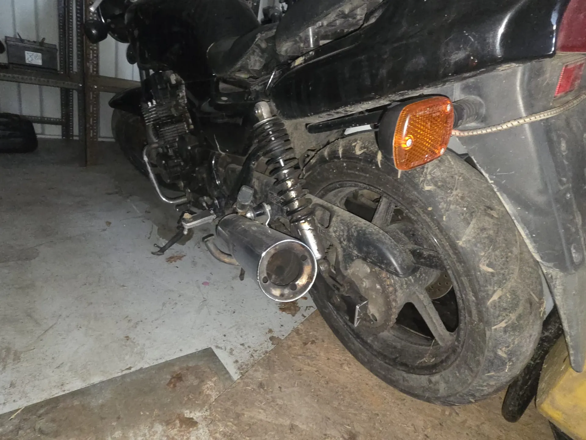 Czarny motocykl widziany od tyłu, z brudnym kołem tylnym, metalowym wydechem i pomarańczowym kierunkowskazem. Motocykl znajduje się w pomieszczeniu z betonową podłogą i metalową półką w tle. Na półce widoczny jest nieczytelny przedmiot. Całość pokryta jest kurzem i śladami brudu.