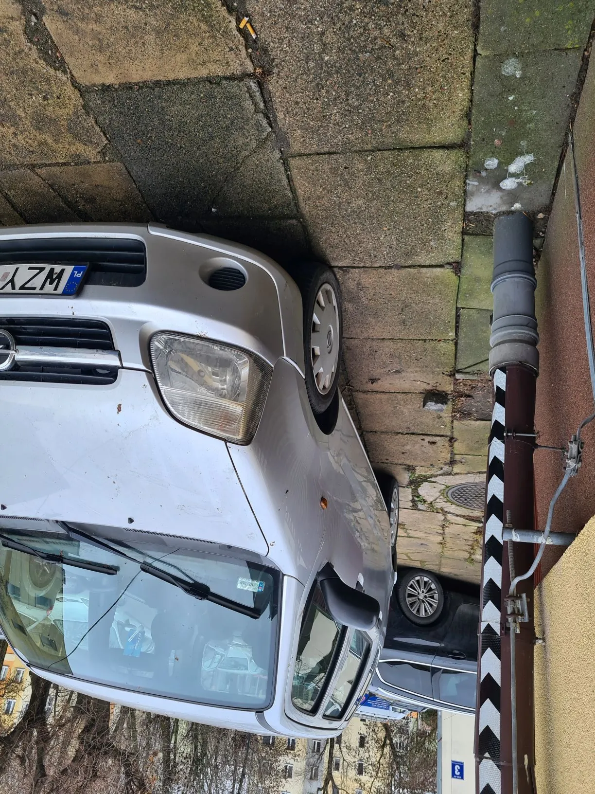 Srebrny samochód zaparkowany na ulicy, widoczna pionowa tablica rejestracyjna z napisem WZY. Przed samochodem bariera z czarno-białymi poziomymi pasami. Na chodniku leży niedopałek papierosa. W tle inne zaparkowane samochody, budynki i drzewa bez liści.