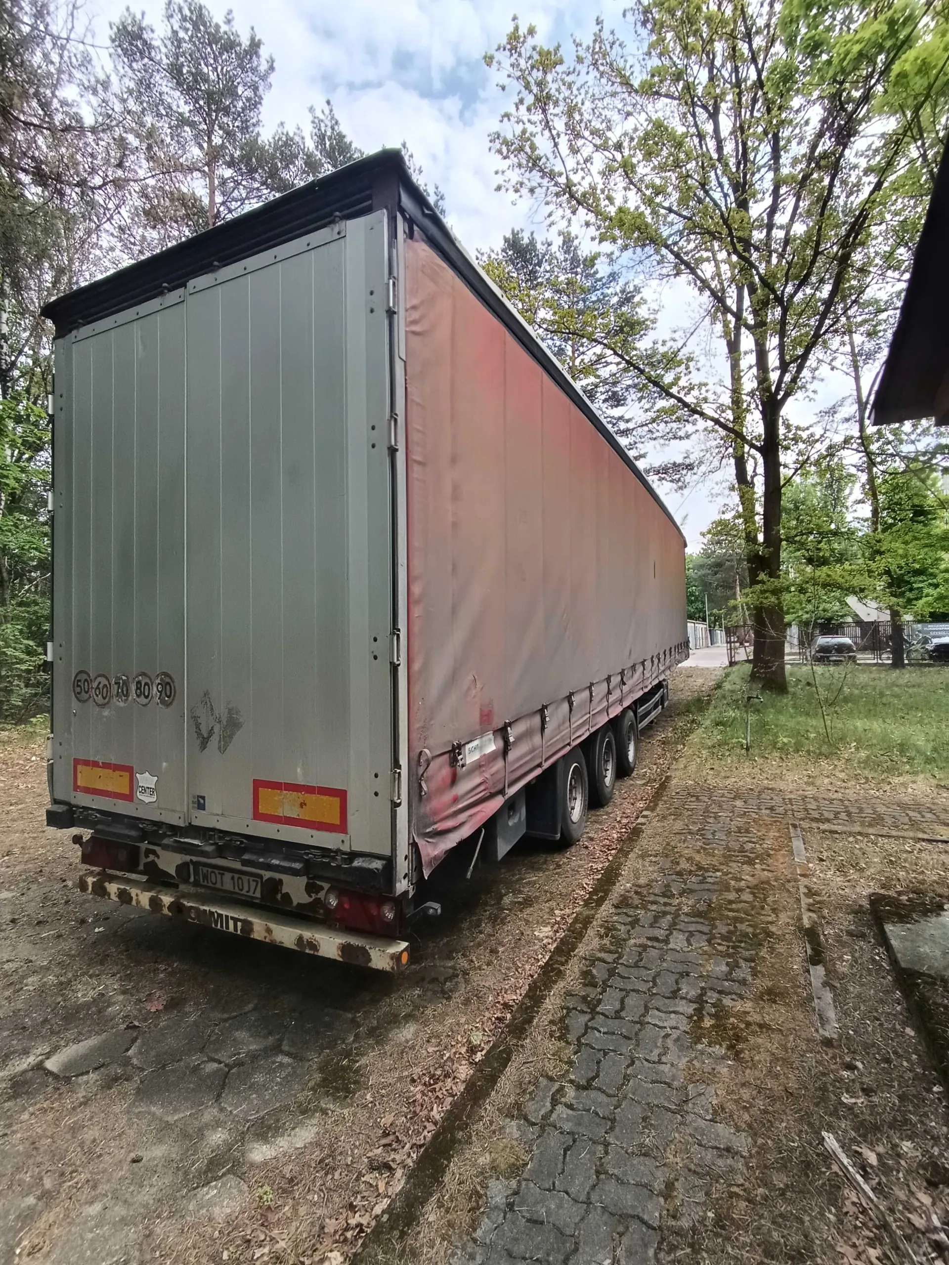 Duży naczepa ciężarowa z tyłu, o szarej metalowej ścianie i brązowym pokryciu bocznym. Na tylnej ścianie widoczne są okrągłe oznaczenia prędkości: 50, 60, 70, 80, 90, czerwone i żółte reflektory oraz tablica rejestracyjna WOT 10J7. Naczepa stoi na brukowanej drodze otoczonej drzewami i trawą. W tle widoczne są drzewa, niebo z chmurami oraz fragment budynku po prawej stronie.