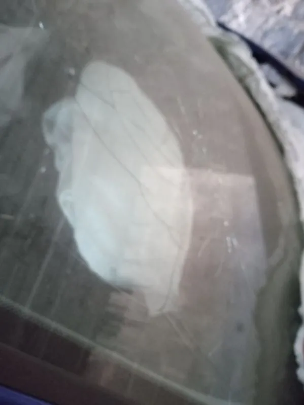 Zdjęcie przedstawia pęknięte szkło, prawdopodobnie przednią szybę samochodu. W centrum widoczne jest duże pęknięcie z rozchodzącymi się od niego mniejszymi rysami. Szkło jest zabrudzone, a w tle rozmyte elementy, w tym fragment materiału po prawej stronie. Brak czytelnego tekstu.