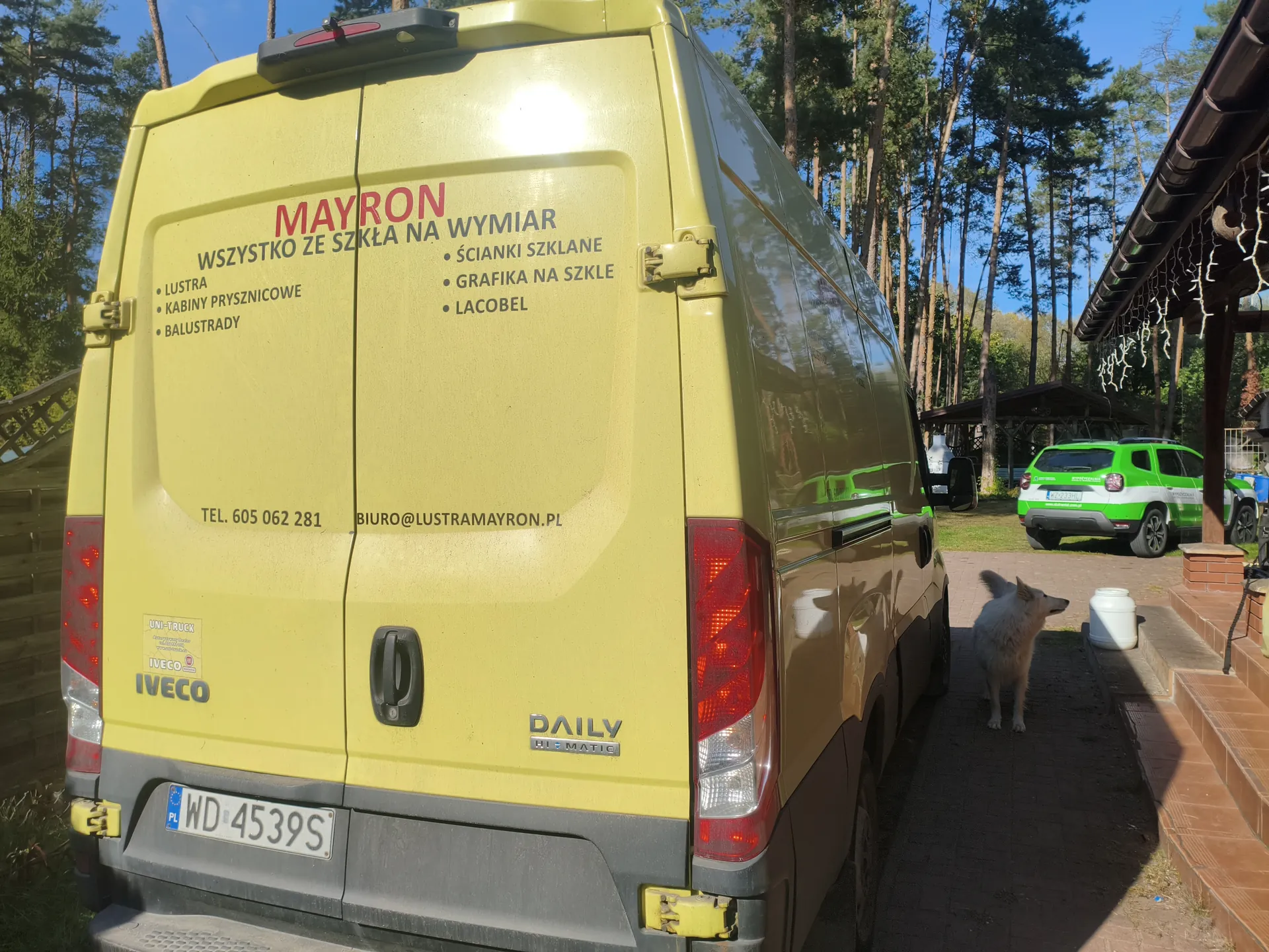 Żółty van IVECO Daily z napisem "MAYRON" i "WSZYSTKO ZE SZKŁA NA WYMIAR", usługami: lustra, kabiny prysznicowe, balustrady, ścianki szklane, grafika na szkle, lacobel. Dane kontaktowe: tel. 605 062 281, biuro@lustramayron.pl, tablica WD 4539S. Obok biały pies, na tle zielony samochód i las.