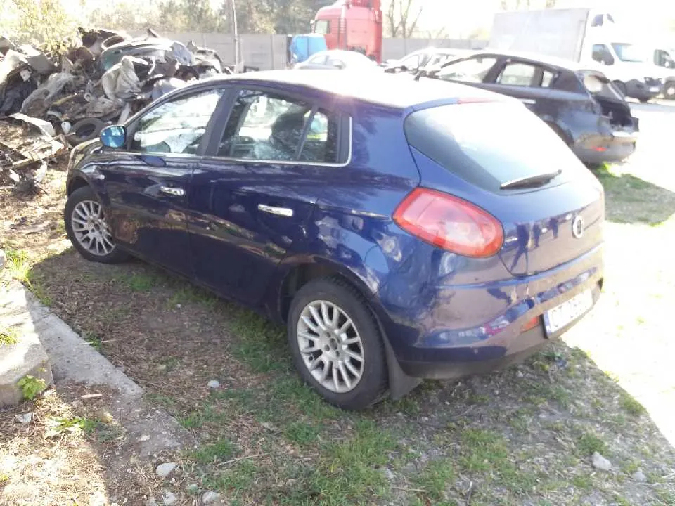 Ciemnoniebieski samochód hatchback zaparkowany na trawie i żwirze, widok od tyłu. W tle widoczne uszkodzone pojazdy, sterta złomu, czerwona ciężarówka oraz biały van. Na tylnej części samochodu umieszczona tablica rejestracyjna z częściowo widocznym numerem.