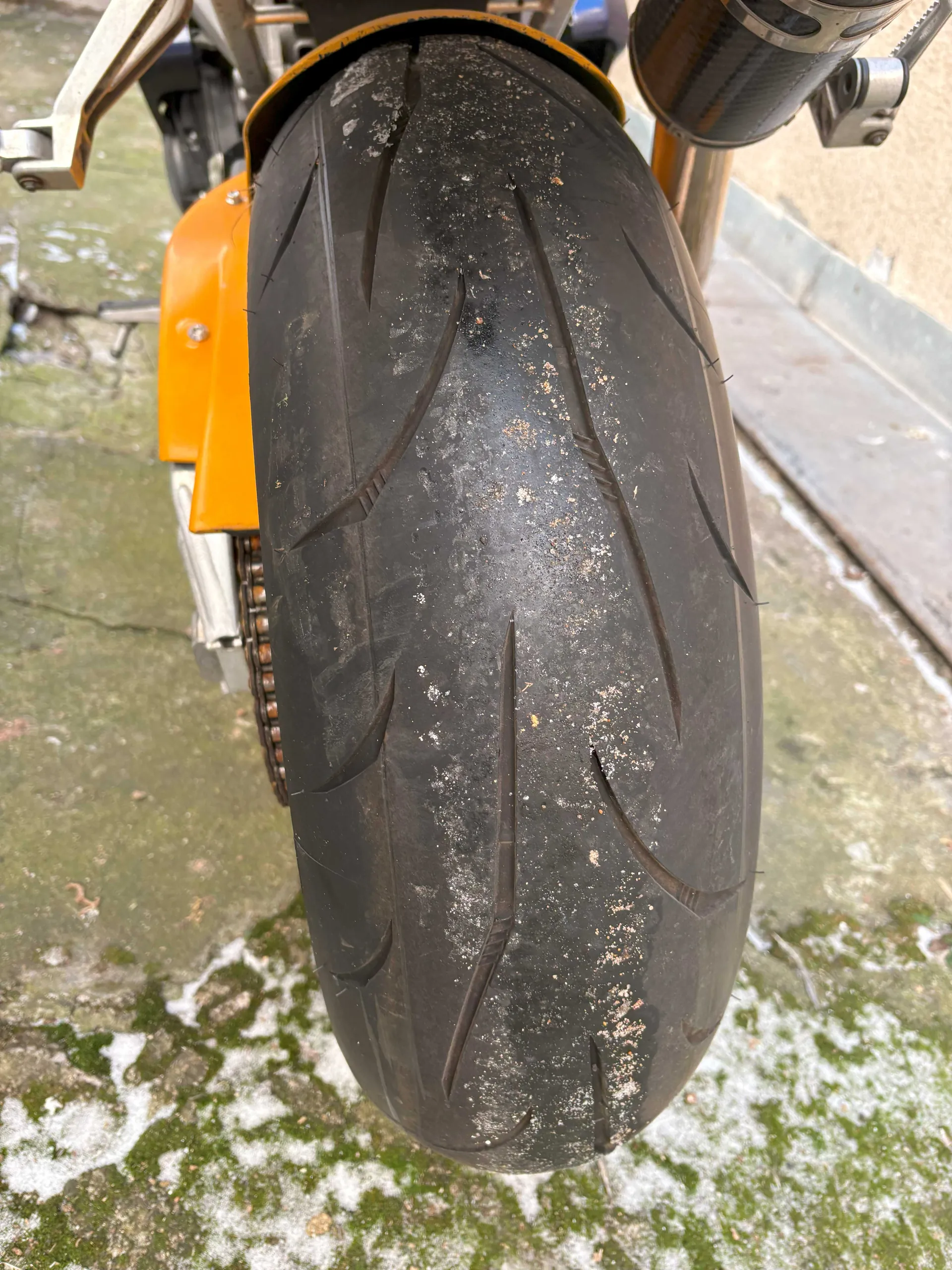 Zbliżenie na czarną oponę motocykla z widocznymi śladami brudu i drobnych cząstek. Opona ma charakterystyczne rysy i wzór gładzi. Po lewej stronie widoczna pomarańczowa część motocykla oraz łańcuch napędowy. Tło stanowi betonowa powierzchnia z plamami mchu i resztkami śniegu.