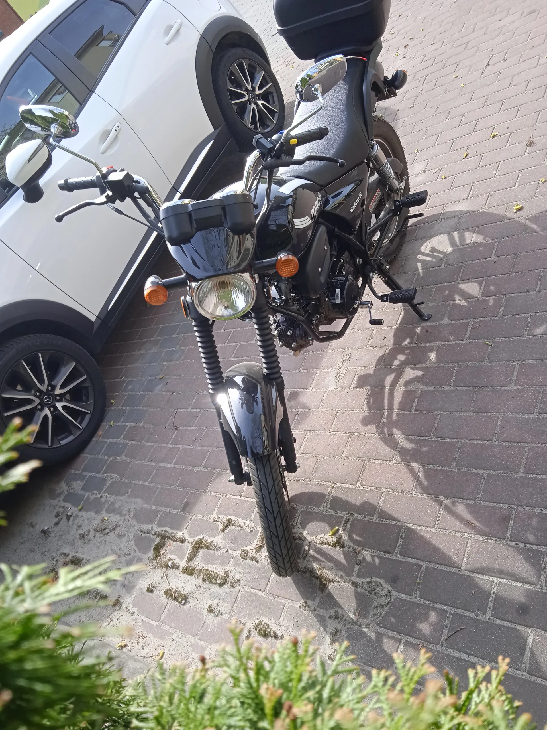 Czarny motocykl z reflektorem, pomarańczowymi kierunkowskazami i lusterkami, zaparkowany na brukowanej powierzchni obok białego samochodu. W tle widoczne są zielone krzewy.