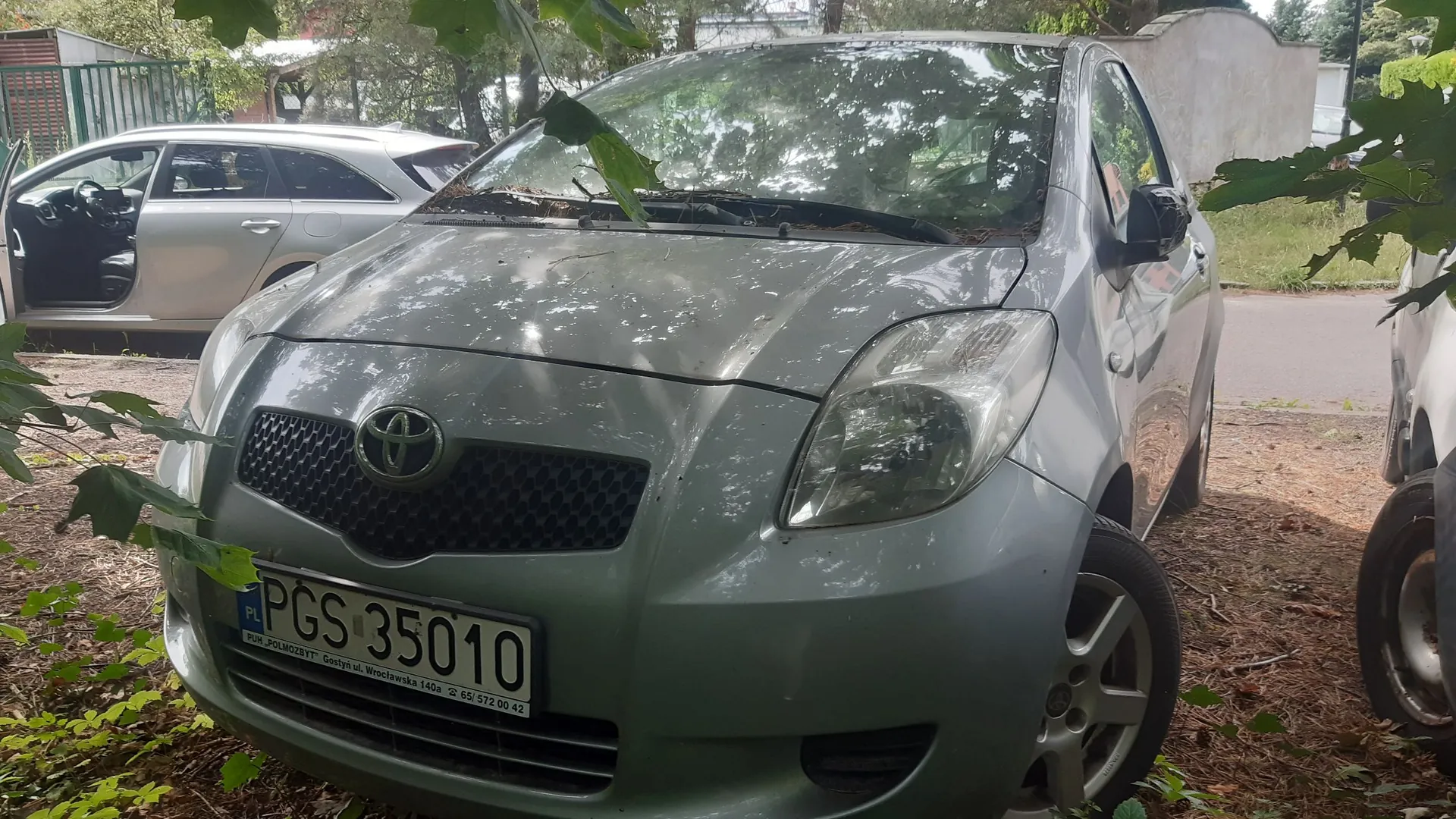 Srebrny samochód marki Toyota zaparkowany na nieutwardzonym terenie w otoczeniu drzew. Widoczny jest przód pojazdu z tablicą rejestracyjną o numerze PGS 35010. W tle po lewej stronie znajduje się inny, srebrny samochód z otwartymi drzwiami.