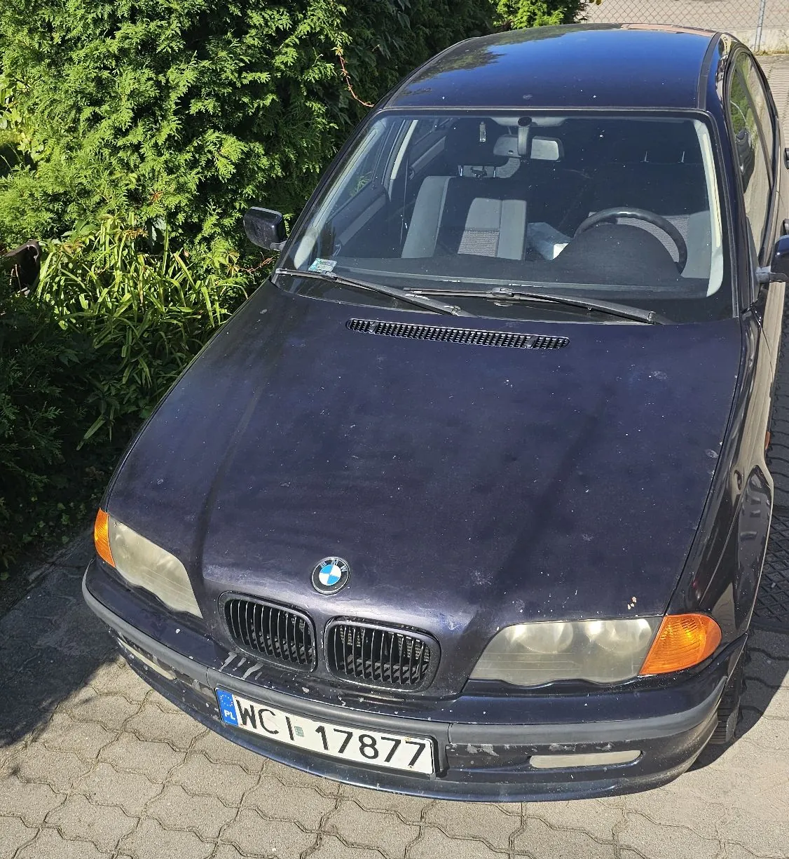 Granatowy samochód marki BMW zaparkowany na kostce brukowej przed zielonym żywopłotem. Widoczny jest przód pojazdu z tablicą rejestracyjną o numerze WCI 17877.
