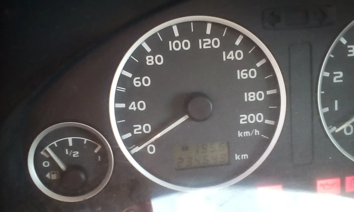 Zbliżenie na deskę rozdzielczą samochodu. Widoczny jest główny prędkościomierz z podziałką od 0 do 200 km/h, wskazówka wskazuje wartość bliską 0. Poniżej znajduje się cyfrowy wyświetlacz z liczbami 195.6 oraz 234545 km. Po lewej stronie widoczny jest mniejszy wskaźnik poziomu paliwa z oznaczeniem 1/2.