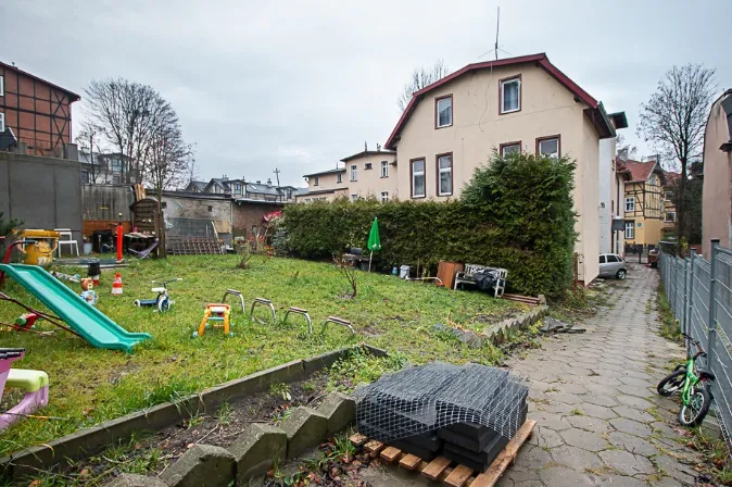 Podwórko z trawnikiem i placem zabaw, na którym znajduje się zielona zjeżdżalnia, małe plastikowe autko oraz kilka metalowych konstrukcji przypominających niskie ławki lub drabinki. Po prawej stronie biegnie ścieżka z kostki brukowej, obok której stoi rower. W tle widoczne są budynki mieszkalne, w tym dom o jasnej elewacji z brązowym dachem oraz budynek z elementami konstrukcji szkieletowej. Teren oddzielony jest od sąsiedztwa wysokim, zielonym żywopłotem.