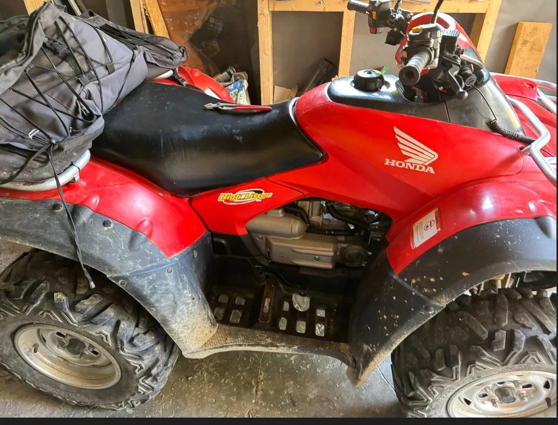 Czerwony quad marki Honda z czarnym siedzeniem i błotnikami. Na tylnej części pojazdu znajduje się przymocowana czarna torba transportowa. Na boku obudowy widoczne jest białe logo Hondy oraz napis 'High Rider'. Pojazd stoi w pomieszczeniu przypominającym garaż lub warsztat.
