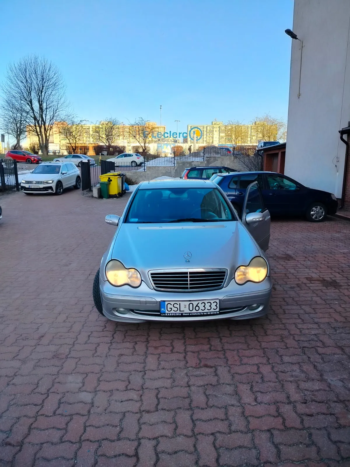 Srebrny samochód marki Mercedes-Benz z przodem skierowanym do przodu, z włączonymi reflektorami i otwartą drzwią po stronie kierowcy. Tablica rejestracyjna z napisem GSL 06333. W tle widoczne są inne zaparkowane pojazdy: biały SUV, ciemnoniebieski samochód i czerwony samochód. W oddali budynek z napisem E.Leclerc, drzewa bez liści, żółty i zielony pojemnik na śmieci oraz budynek po prawej stronie. Niebo jest jasne, a podłoga wyłożona czerwoną kostką brukową.