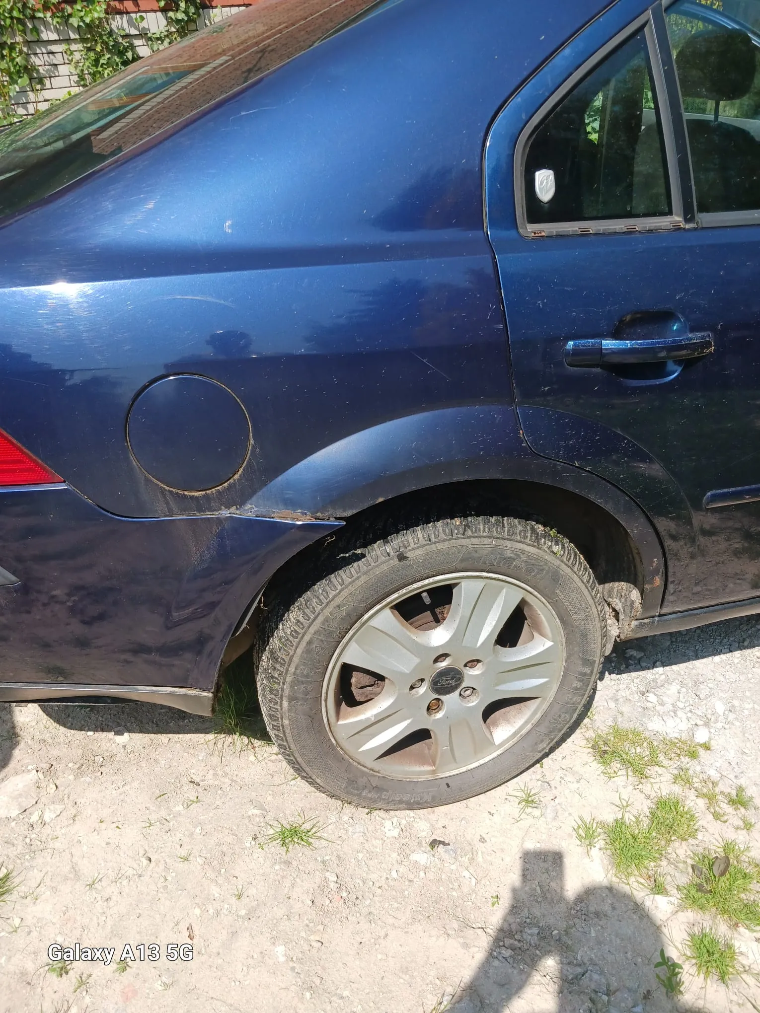 Niebieski samochód osobowy, widok z boku na tylną część. Widoczne są tylne koło z srebrną felgą z logo Ford, zamknięty korek baku, fragment tylnej lampy i drzwi z uchwytem. Samochód stoi na podłożu z żwiru i trawą. Na dole zdjęcia widoczny napis "Galaxy A13 5G". W tle fragment ceglanej ściany z zielonymi pnączami. Na powierzchni samochodu widoczne są ślady brudu.