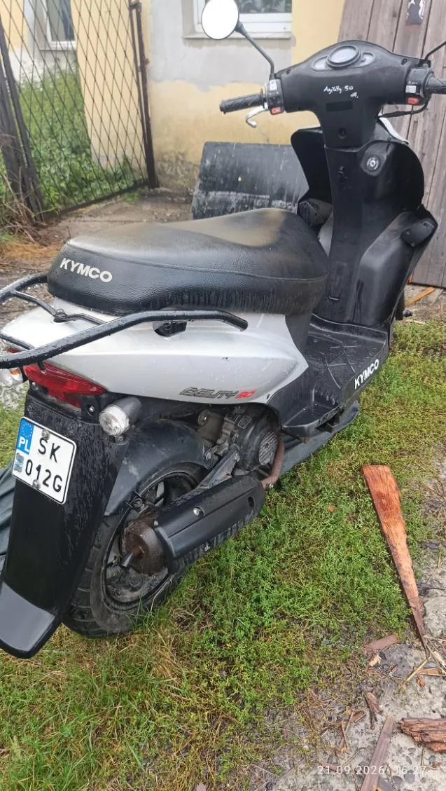 Srebrno-czarny skuter marki Kymco modelu Beauty stojący na trawie. Na siedzeniu widnieje napis KYMCO, a na boku obudowy napis BEAUTY. Pojazd posiada tablicę rejestracyjną z oznaczeniem PL SK 2012G. W prawym dolnym rogu zdjęcia znajduje się data 21.09.2023 16:27.