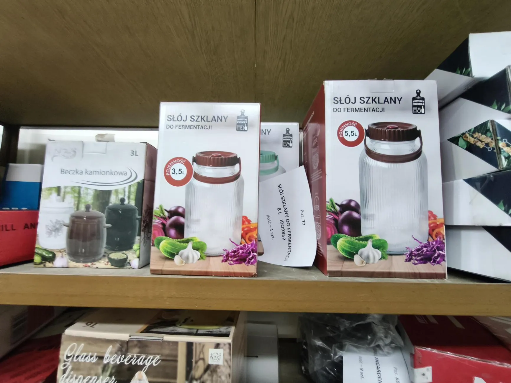 Zdjęcie produktów na drewnianej półce sklepowej. Widoczne są trzy opakowania: po lewej białe pudełko z napisem 'Beczka kamionkowa 3L' i zdjęciem trzech ceramicznych naczyń; w środku pudełko z napisem 'SŁÓJ SZKLANY DO FERMENTACJI' o pojemności '3,5L' z obrazkiem szklanego słoja z brązową pokrywką; po prawej większe pudełko z napisem 'SŁÓJ SZKLANY DO FERMENTACJI' o pojemności '5,5L', również z obrazkiem szklanego słoja z brązową pokrywką. Na opakowaniach słoików widoczne są dodatkowo zdjęcia warzyw: czerwonej kapusty, czosnku i bakłażana.