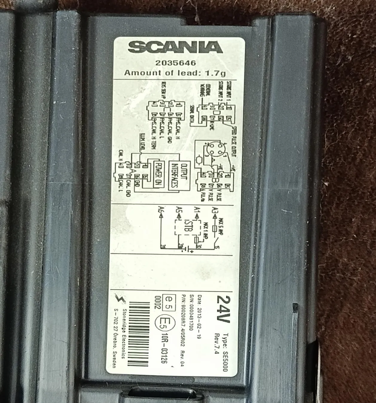 Czarna obudowa z białą etykietą zawierającą tekst i schematy. Na etykiecie widnieje napis 'SCANIA', numer '2035646', informacja 'Amount of lead: 1.7g', schematy elektryczne, oznaczenia '24V', 'Type: SE5000', 'Rev 7.4', numery seryjne 'S/N: 0000481700', 'P/N: 90020887-405802 Rev 04', kod kreskowy oraz informacja o producencie 'Stoneridge Electronics S-702 27 Örebro, Sweden'. Tło po prawej stronie etykiety to brązowa, włóknista powierzchnia.
