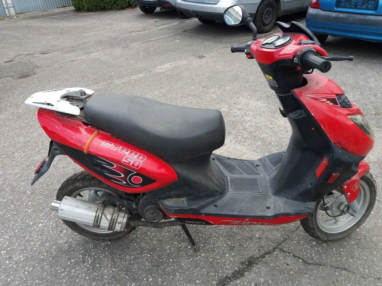 Czerwono-czarny skuter z napisem "Speed 50" na boku, czarnym siedzeniem i pustym białym uchwytem na tablicę rejestracyjną. Skuter stoi na asfaltowym parkingu z widocznymi pęknięciami. W tle znajdują się częściowo widoczne pojazdy: srebrny i niebieski samochód, a także zieleń i metalowy płot. Skuter wyposażony jest w lusterka, dwa okrągłe wskaźniki na kierownicy oraz srebrny tłumik.