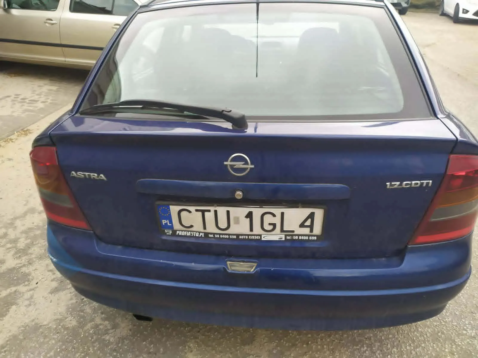 Tył niebieskiego samochodu Opel Astra z widocznym logo marki, napisem "ASTRA" po lewej stronie i "1.7 CDTI" po prawej. Tablica rejestracyjna z polskim kodem "PL" i numerem "CTU 1GL4". Poniżej tablicy napisy: "PROFIXITO.PL", numer telefonu "tel. 58 6400 698" oraz "AUTO CZĘŚCI". W tle częściowo widoczny srebrny samochód i asfaltowy parking.