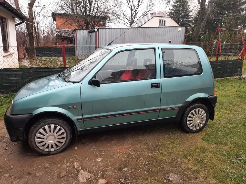 Mały samochód osobowy w kolorze turkusowym zaparkowany na podwórku. Samochód ma czerwone siedzenia, czarne zderzaki i srebrne felgi. W tle widoczne są domy mieszkalne, metalowy garaż, ogrodzenie z siatką, drzewa bez liści oraz czerwona konstrukcja placu zabaw. Podłoże składa się z trawy i odsłoniętej ziemi.