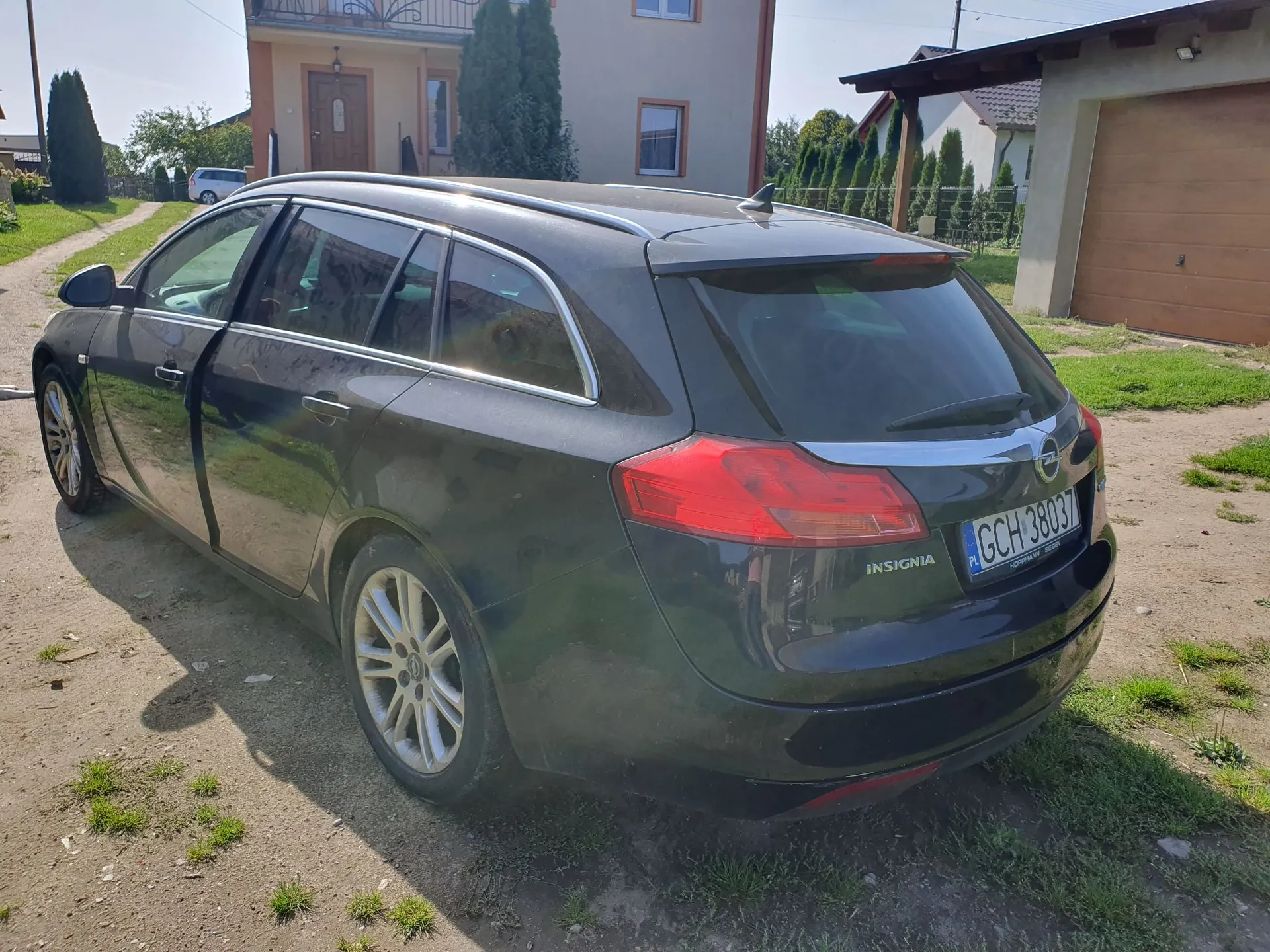 Czarny samochód marki Opel Insignia w nadwoziu kombi, zaparkowany na piaszczystej drodze przed domem. Widoczna jest tylna część pojazdu z tablicą rejestracyjną GCH 38037. W tle znajduje się jasny budynek mieszkalny, zielone krzewy oraz garaż z brązową bramą.