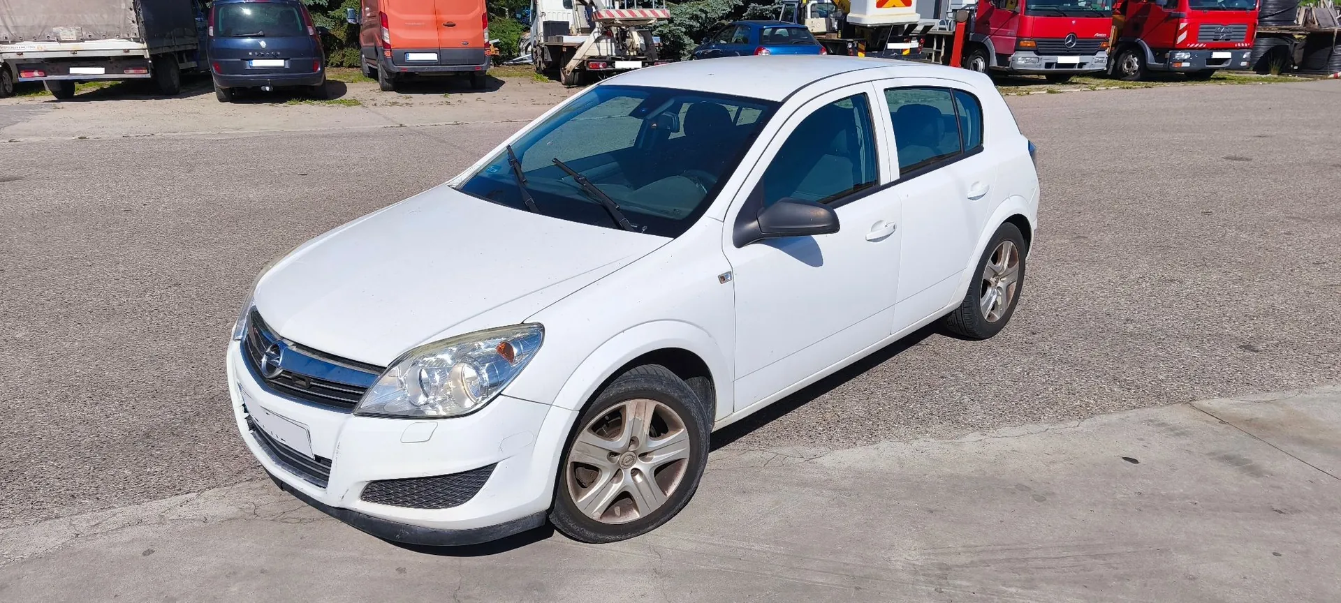 Biały samochód osobowy marki Opel Astra w nadwoziu hatchback, zaparkowany na asfaltowym placu. Pojazd jest widoczny z przodu i z boku. W tle znajdują się inne pojazdy, w tym czerwone ciężarówki i pomarańczowy furgon.