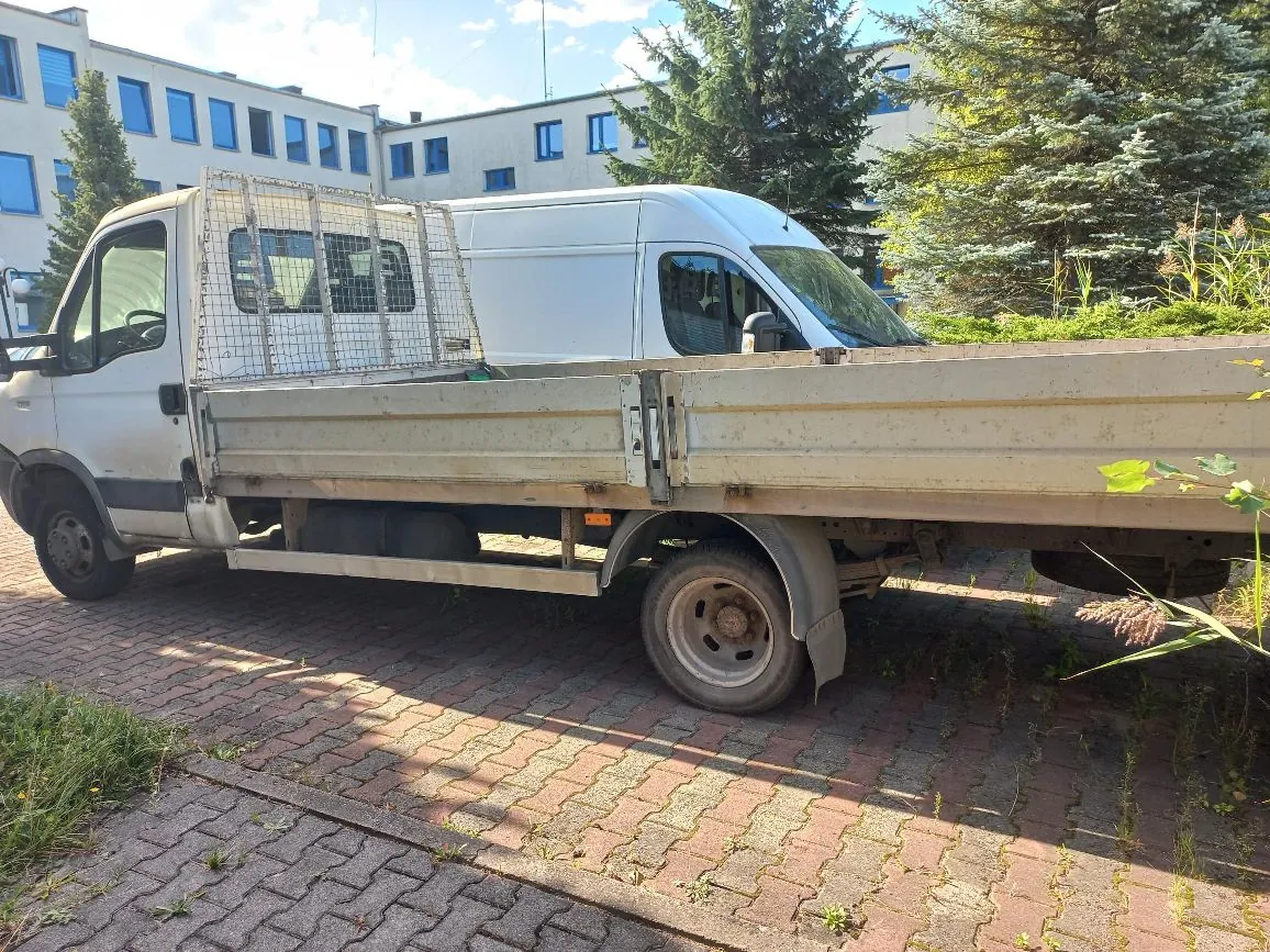 Biały samochód dostawczy z otwartą platformą ładunkową, zaparkowany na kostce brukowej. Z tyłu kabiny znajduje się metalowa kratka ochronna. W tle widoczny jest jasny budynek z niebieskimi ramami okien oraz zielone drzewa i krzewy.