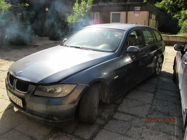 Ciemnoszary samochód osobowy marki BMW w nadwoziu kombi, zaparkowany na kostce brukowej. Pojazd jest widoczny z przodu i z boku, z widoczną tablicą rejestracyjną o numerze LJ 68232. W tle znajduje się budynek z jasnymi ścianami oraz zieleń w postaci krzewów i drzew. W prawym dolnym rogu zdjęcia znajduje się pomarańczowa data: 08/09/2026.