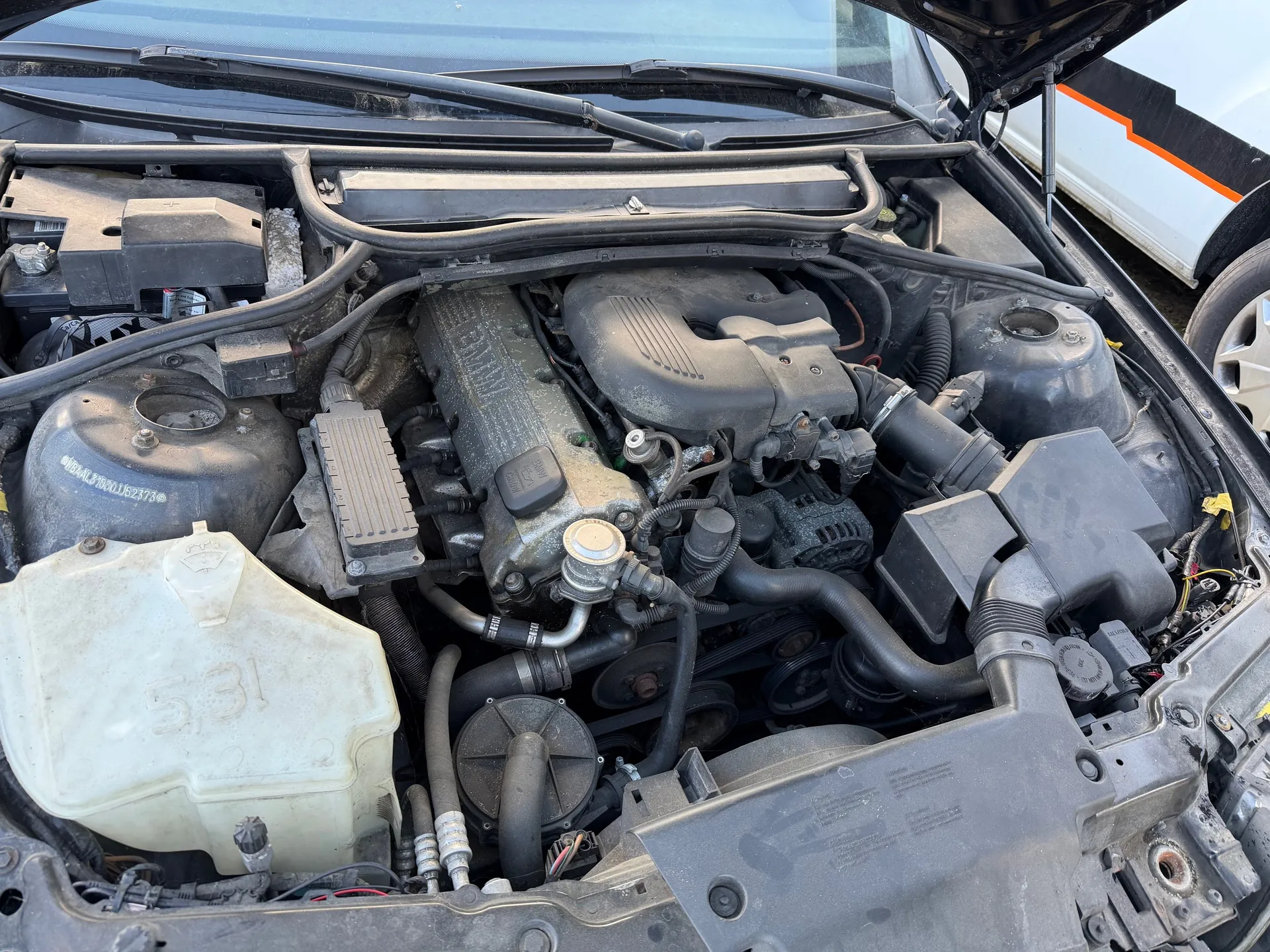 Zdjęcie przedstawia otwarty maskę samochodu z widocznym silnikiem. W centralnej części znajduje się blok silnika z czarnymi przewodami i elementami. Po lewej stronie widoczny jest biały zbiornik z napisem "5.3L" oraz czarny zbiornik z częściowo czytelnym numerem "WBA...52373". W tle, po prawej stronie, fragment innego pojazdu z białą karoserią i pomarańczową listwą. Całość ukazana jest z góry, z silnikiem i elementami układu chłodzenia.