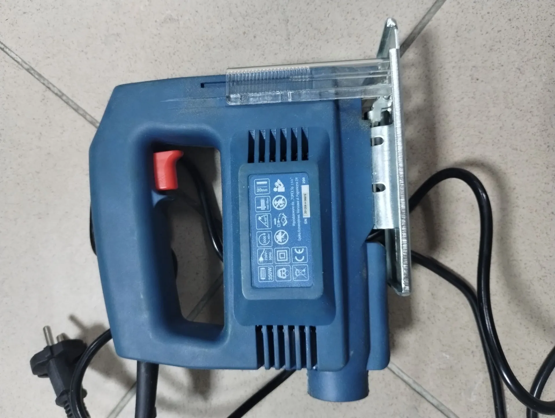 Niebieska wyrzynarka elektryczna leżąca na jasnej, kafelkowej powierzchni. Urządzenie posiada uchwyt z czerwonym przełącznikiem oraz metalową stopę. Na boku obudowy znajduje się tabliczka znamionowa z tekstem i symbolami, w tym widoczne napisy: '350W', 'CE' oraz 'SNA 2013-00015'. Z urządzenia wychodzi czarny kabel z wtyczką elektryczną.