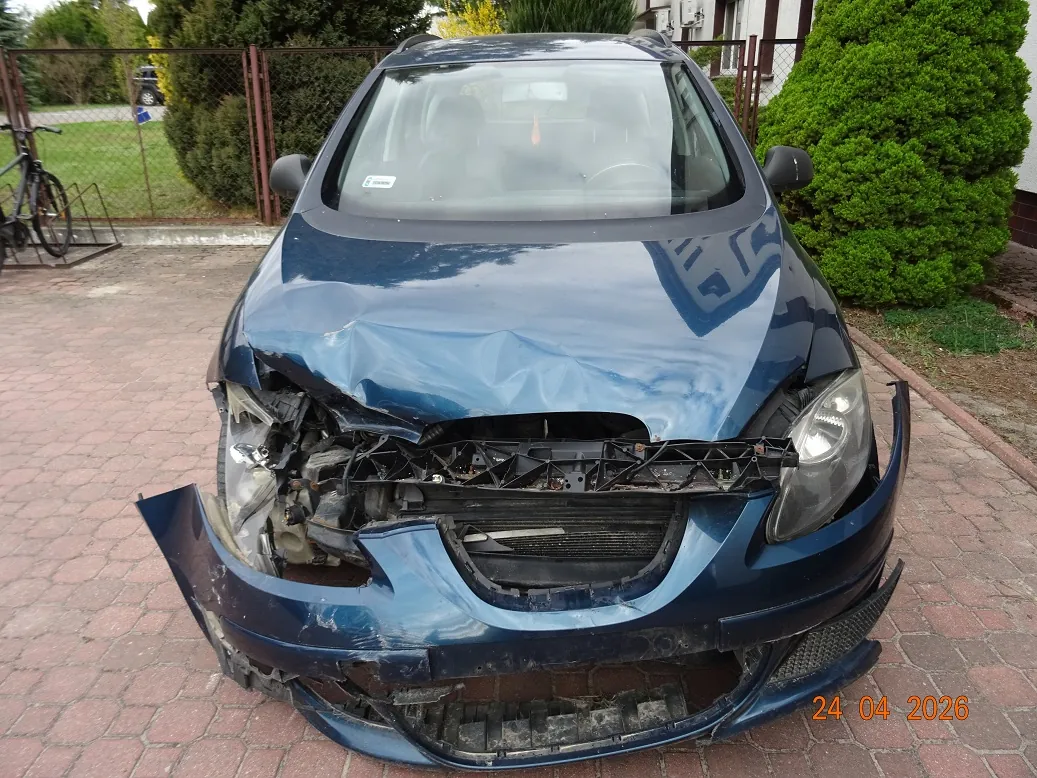Zdjęcie przodu ciemnoniebieskiego samochodu osobowego z poważnymi uszkodzeniami po kolizji. Maską jest wgnieciona, przedni zderzak jest częściowo oderwany i pęknięty, a elementy konstrukcyjne pod maską są widoczne i zniekształcone. Lewy reflektor jest rozbity. Samochód stoi na kostce brukowej na tle zielonych krzewów i ogrodzenia. W prawym dolnym rogu znajduje się pomarańczowa data: 24 04 2026.