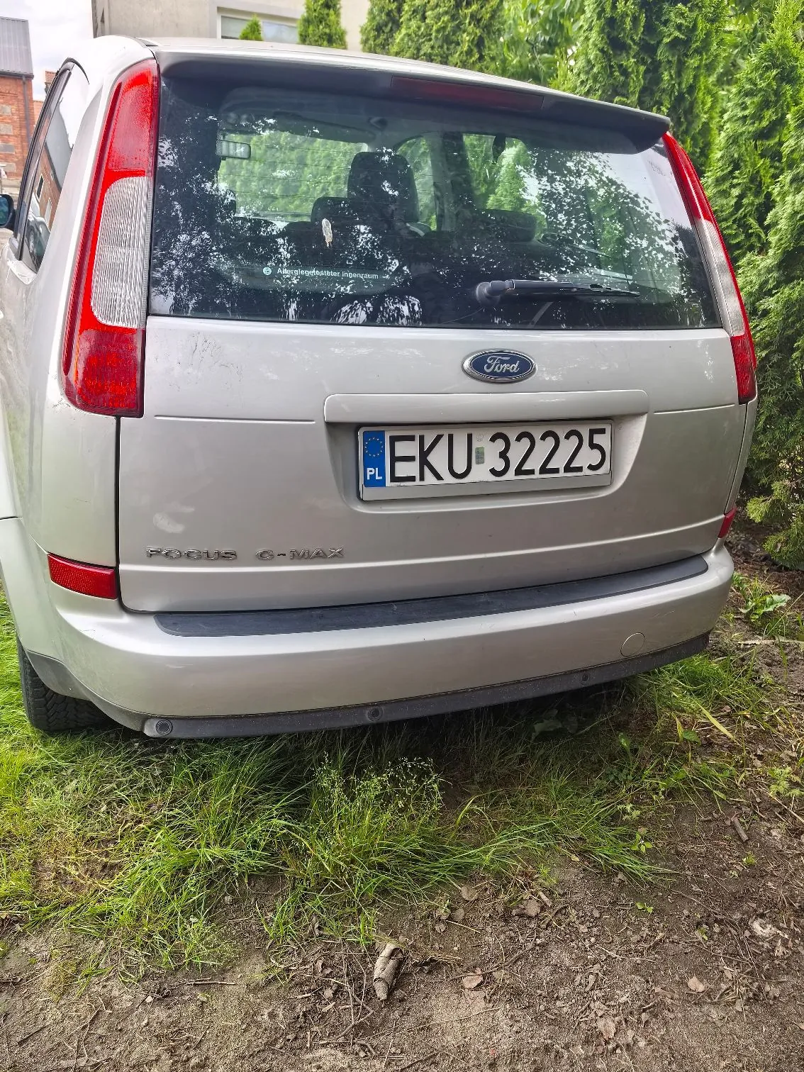 Tylny widok srebrnego samochodu marki Ford Focus C-MAX. Na klapie bagażnika znajduje się logo Ford oraz tablica rejestracyjna o numerze EKU 32225 z oznaczeniem PL. Poniżej tablicy widnieje napis FOCUS C-MAX. Samochód zaparkowany jest na trawiastym podłożu obok zielonego żywopłotu.