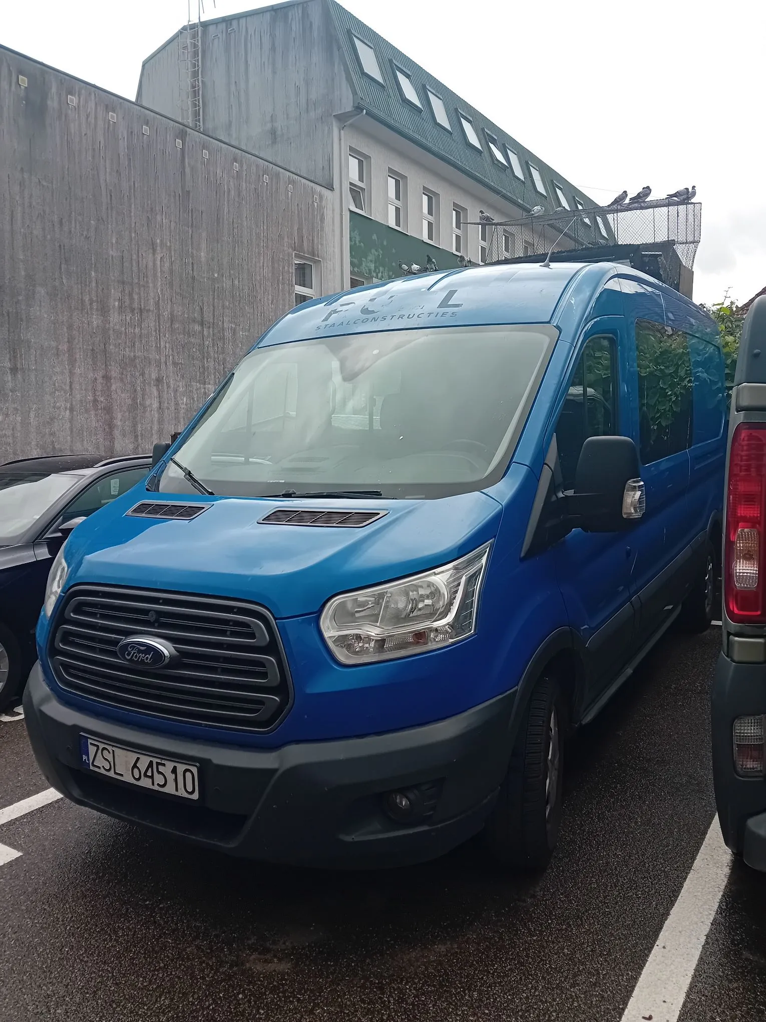 Niebieski furgon marki Ford zaparkowany na parkingu przed szarym budynkiem. Na dachu samochodu znajduje się bagażnik, a na przedniej części nad szybą widnieje napis „F L STEELCONSTRUCTIONS”. Pojazd ma polską tablicę rejestracyjną o numerze ZSL 64510.