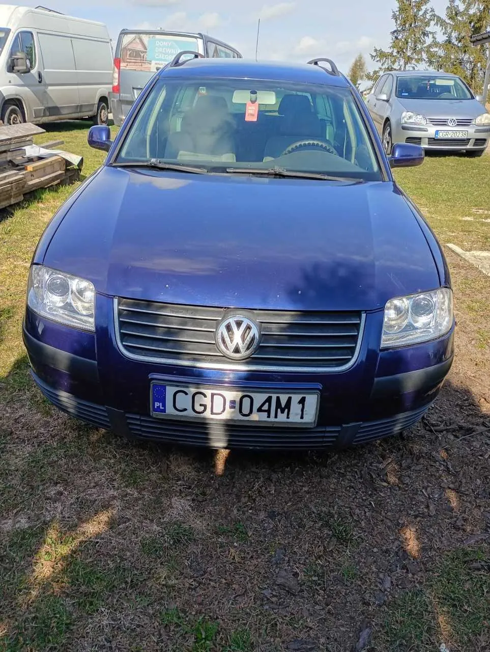 Niebieski samochód osobowy marki Volkswagen z tablicą rejestracyjną CGD-04M1 (PL) na pierwszym planie. Na przedniej szybie widoczny czerwony znaczek. W tle stoją inne pojazdy: srebrny furgon, ciężarówka z napisem "DOMY DREWNIANE" oraz srebrny samochód osobowy. Scena rozgrywa się na trawiastym obszarze z drzewami i niebem w tle.